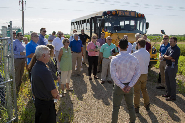 A crowd listening to a man while standing near an electric school bus.